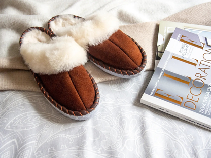 Brown shearling sheepskin mules on a bed next to magazines.