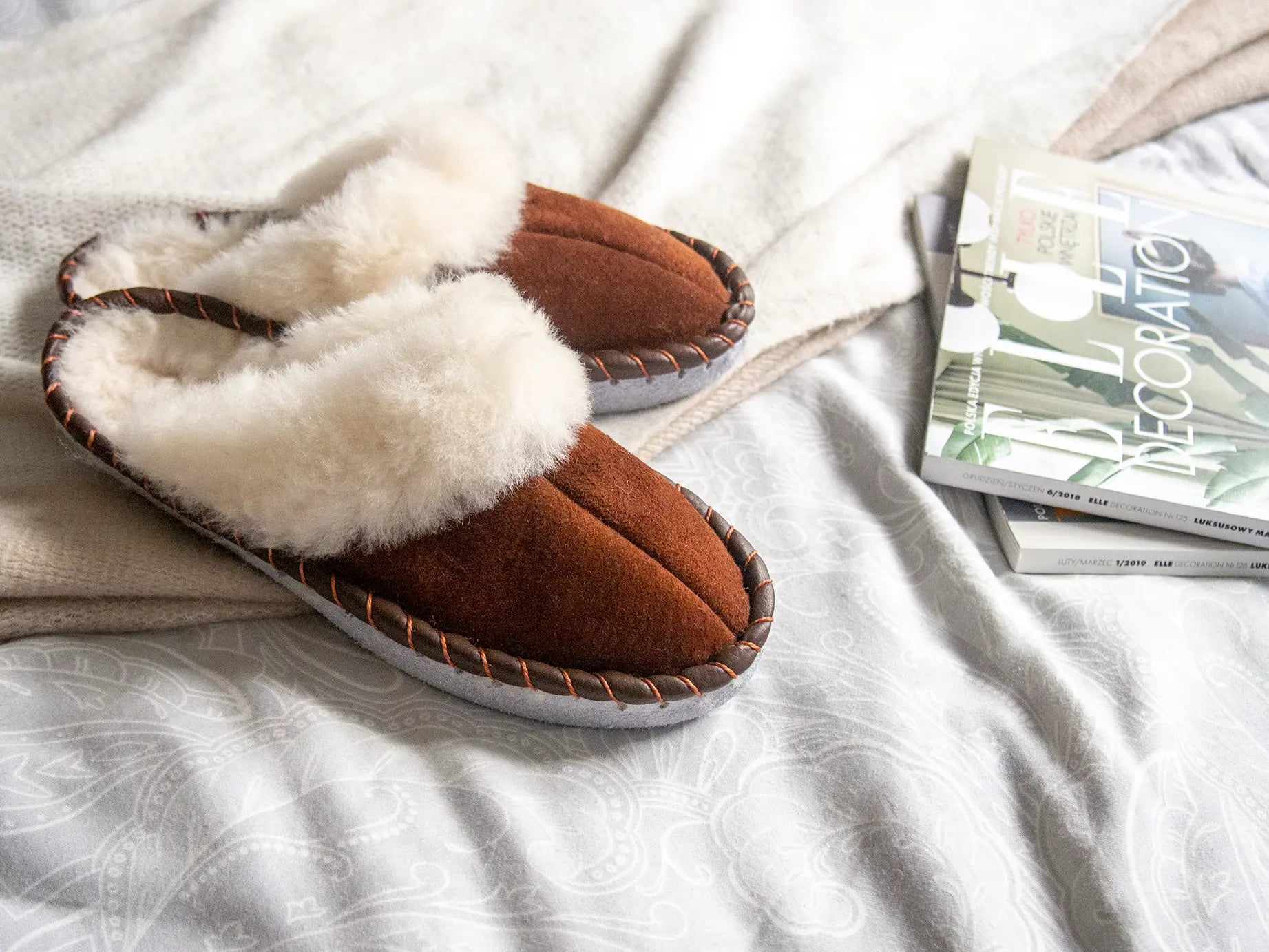 Brown shearling sheepskin mules on a bed with magazines.