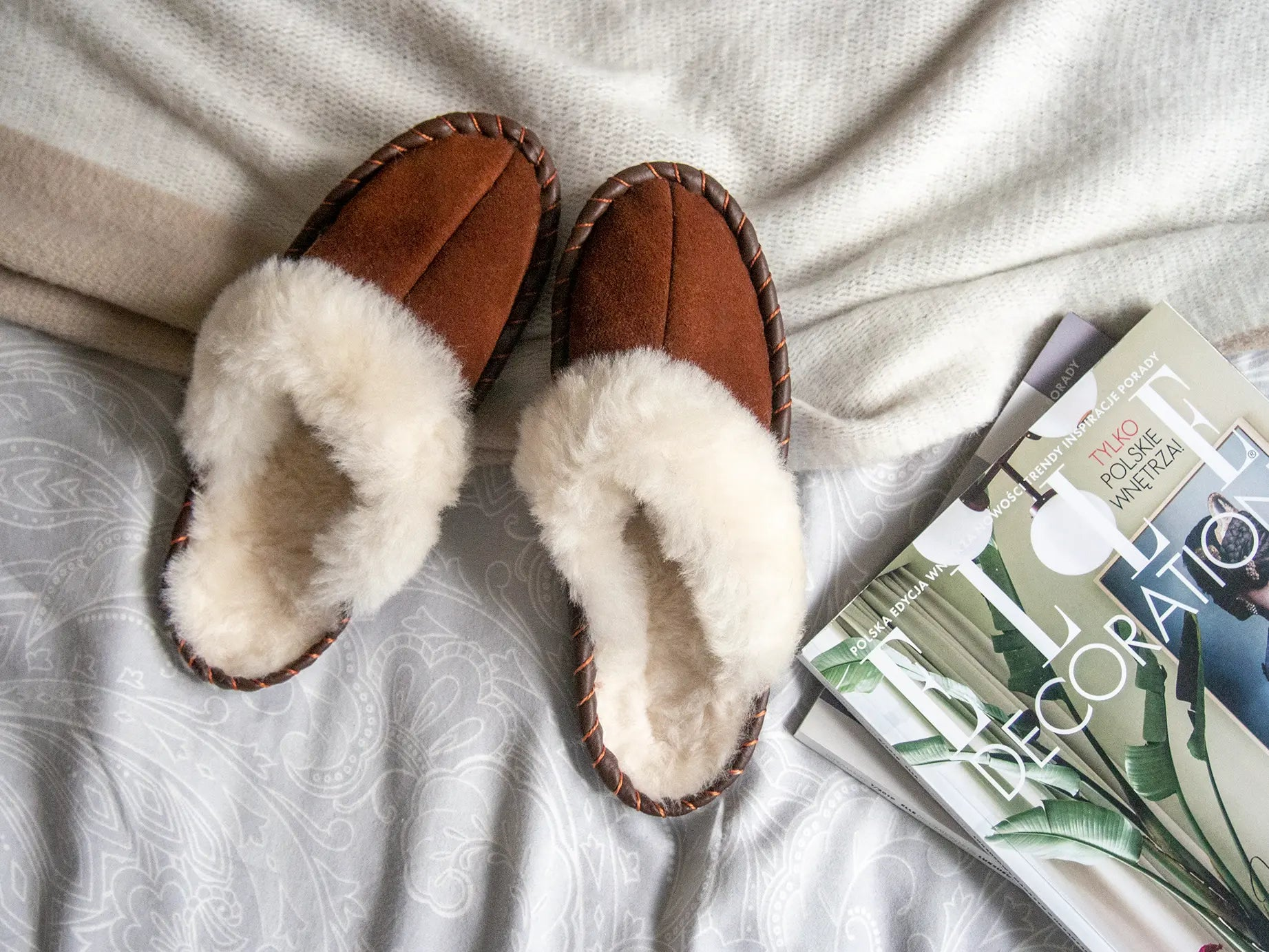 Brown shearling sheepskin mules with natural leather trim on a cozy blanket beside magazines.